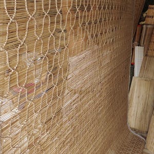 May include: A large, rectangular bamboo mat featuring thin, light brown horizontal slats held together by a repeating, wavy white cord pattern. The mat has a thick, light-colored fabric border along the edges and a smooth, natural finish.