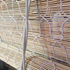 May include: Close-up of a bamboo window shade with a woven design and a light-colored rope for raising and lowering. The shade has a wooden mechanism at the top and a white fabric border.