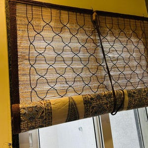 May include: A bamboo roll-up window shade with a decorative patterned fabric at the bottom. The shade has a dark brown border and a black crisscross pattern. The fabric features a beige background with blue and brown designs.