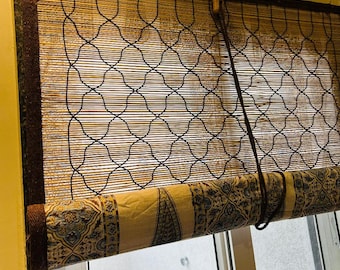 Pode incluir: Uma persiana de bambu enrolável com um tecido estampado na parte inferior. A persiana tem uma borda marrom escura e um design de treliça preto. O tecido apresenta um fundo bege com desenhos azuis e marrons.