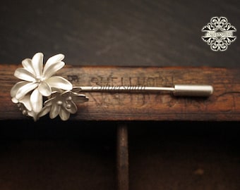 Puede incluir: Un broche de plata con forma de flor con un alfiler en la parte posterior. El broche está hecho de tres flores con pétalos redondeados y algunas pequeñas cuentas redondas en el centro.