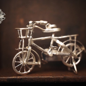 Puede incluir: Una bicicleta en miniatura de plata con una cesta y un portaequipajes trasero. La bicicleta está sobre una superficie de madera.
