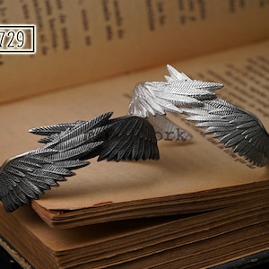 May include: Two silver wing-shaped bracelets, one with a darker finish than the other, are displayed on a stack of old books. The bracelets are made of metal and have a detailed design of feathers.