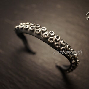 Könnte beinhalten: Ein silbernes Armband mit einem Kraken-Tentakel-Design. Das Armband ist aus Metall und hat eine strukturierte Oberfläche. Der Tentakel ist mit kleinen Kreisen verziert.