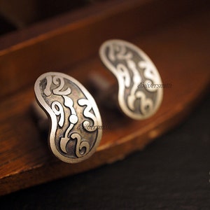 May include: A pair of silver cufflinks with a kidney bean shape and intricate designs featuring numbers. The cufflinks are on a wooden surface.