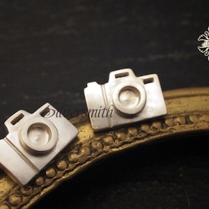May include: Two silver camera shaped earrings on a gold frame.