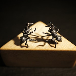 May include: Two black ant-shaped earrings on a wooden surface. The earrings are made of metal and have a detailed design.