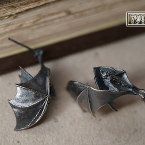 Op de afbeelding: Een paar zilveren vleermuis oorbellen met gedetailleerde vleugels en een vleermuis die aan elke vleugel hangt. De oorbellen hangen aan een houten oppervlak.