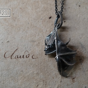 May include: A silver bat pendant necklace with a detailed design. The bat is hanging upside down with its wings spread out. The necklace is on a silver chain.