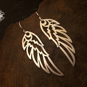 May include: A pair of silver-colored wing-shaped earrings with intricate cut-out designs. The earrings are displayed on a dark wooden surface, showcasing their detailed craftsmanship. The earrings have a hook closure.