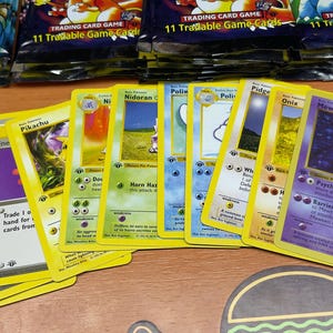 May include: A collection of Pok&eacute;mon trading cards, featuring characters like Pikachu and Mewtwo, with yellow borders and various illustrations. Packs of trading cards are visible in the background.