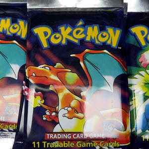 May include: Sealed Pok&eacute;mon trading card game packs. The packs are dark blue with the Pok&eacute;mon logo in yellow. The front of the pack features a Charizard illustration. The text "Trading Card Game" and "11 Tradable Game Cards" are also visible.