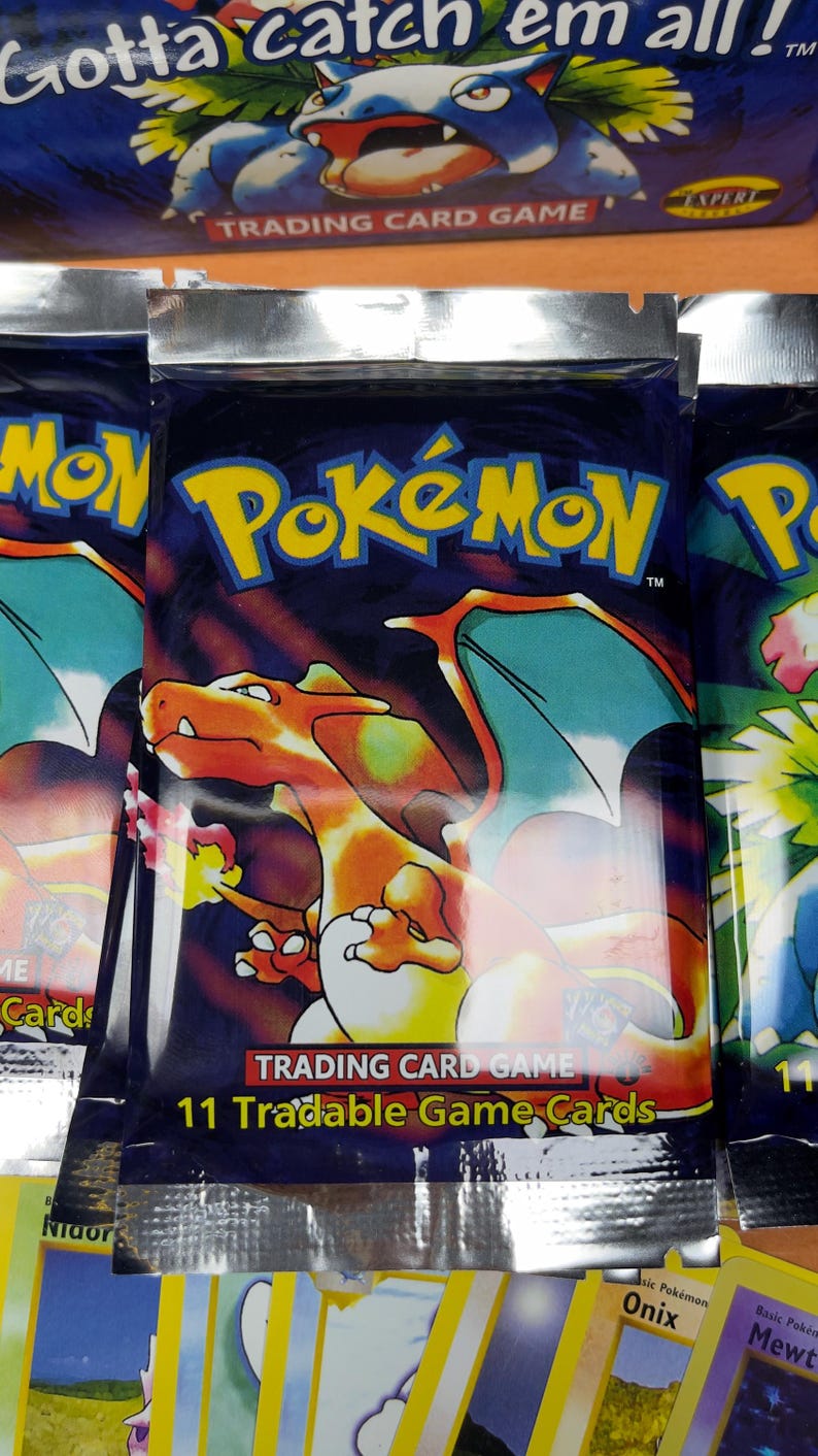 May include: Sealed Pok&eacute;mon trading card game packs. The packs are dark blue with the Pok&eacute;mon logo in yellow. The front of the pack features a Charizard illustration. The text "Trading Card Game" and "11 Tradable Game Cards" are also visible.