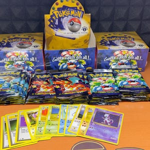 Può includere: Una collezione di oggetti del gioco di carte collezionabili Pokémon. L'immagine mostra buste, scatole e singole carte non aperte. Le scatole sono gialle e blu, con il logo Pokémon e la scritta "Gotta catch 'em all!".