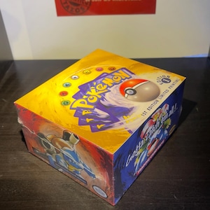 May include: A yellow and blue Pok&eacute;mon trading card game box with the text "Gotta Catch 'Em All!" and "1st Edition Limited Printing." The box features images of Pok&eacute;mon characters and a Pok&eacute; Ball. The box is on a dark surface.