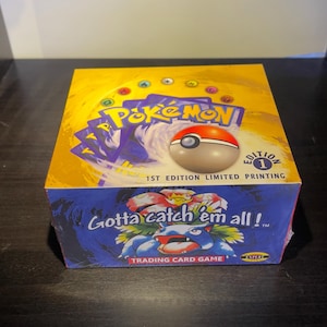 May include: A sealed Pok&eacute;mon trading card game box. The box is yellow and blue with the Pok&eacute;mon logo and a Pok&eacute;ball on top. The text "Gotta catch 'em all!" is visible.