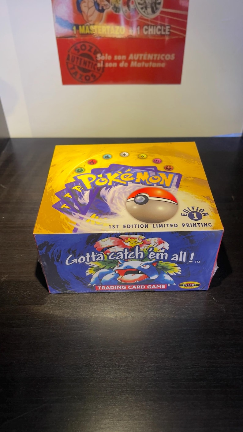 May include: A sealed Pok&eacute;mon trading card game box. The box is yellow and blue with the Pok&eacute;mon logo and a Pok&eacute;ball on top. The text "Gotta catch 'em all!" is visible.