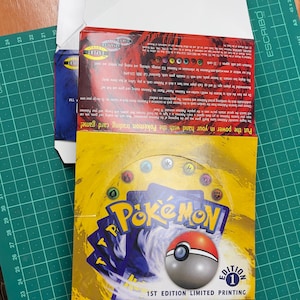 May include: A yellow and blue Pok&eacute;mon trading card game box with the text "Pok&eacute;mon" and a Pok&eacute; Ball graphic. The box is labeled "1ST EDITION LIMITED PRINTING". The box is open, revealing the cards inside.