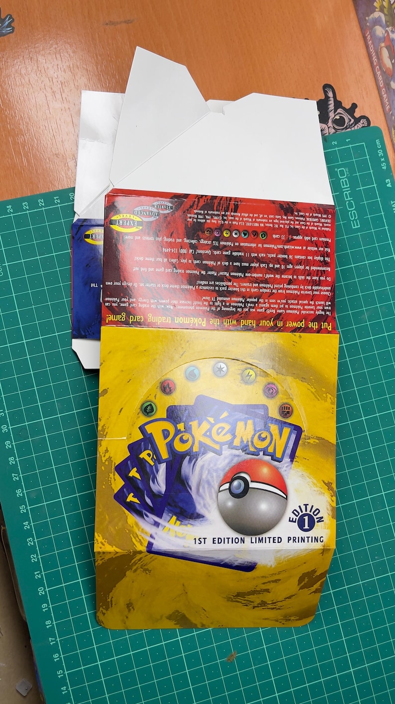 May include: A yellow and blue Pok&eacute;mon trading card game box with the text "Pok&eacute;mon" and a Pok&eacute; Ball graphic. The box is labeled "1ST EDITION LIMITED PRINTING". The box is open, revealing the cards inside.