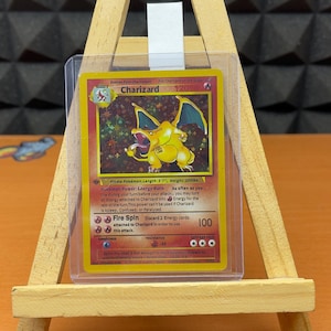 May include: A Charizard Pokémon card, featuring a yellow and orange dragon-like creature with blue wings. The card is in a protective sleeve and displayed on a wooden easel. The card text includes "Fire Spin" and "100".