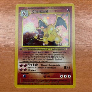 Puede incluir: Una carta coleccionable de Pokémon Charizard. La carta presenta una criatura con forma de dragón amarillo y naranja con alas extendidas. La carta tiene texto que incluye el nombre "Charizard", "Fire Spin" y otra información relacionada con el juego. La carta mide 170 cm de largo y pesa 90 kg.