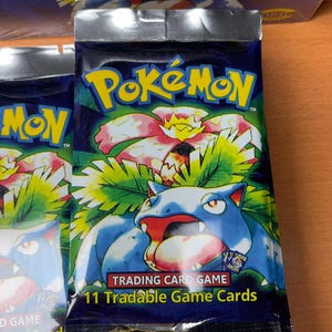 May include: Sealed Pok&eacute;mon trading card game pack. The pack features the Pok&eacute;mon logo in yellow and blue, with a blue and red creature illustration. The text "11 Tradable Game Cards" is visible. The packaging is silver and blue.