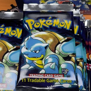 May include: Sealed Pok&eacute;mon trading card packs featuring Blastoise artwork. The packs are silver and blue, with the Pok&eacute;mon logo and text that reads "11 Tradable Game Cards". The box in the background reads "Gotta catch 'em".