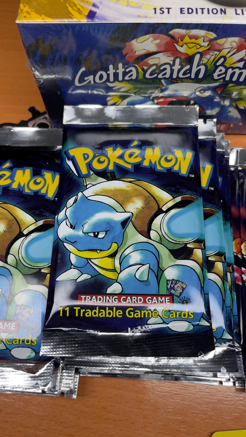 May include: Sealed Pok&eacute;mon trading card packs featuring Blastoise artwork. The packs are silver and blue, with the Pok&eacute;mon logo and text that reads "11 Tradable Game Cards". The box in the background reads "Gotta catch 'em".
