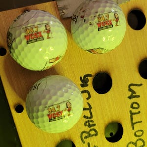 May include: Three white golf balls with dimpled surfaces, each featuring a printed logo that reads 94.7 WCSX Classic Rock with a cartoon character. The balls are positioned on a yellow board with black circular holes and handwritten text.