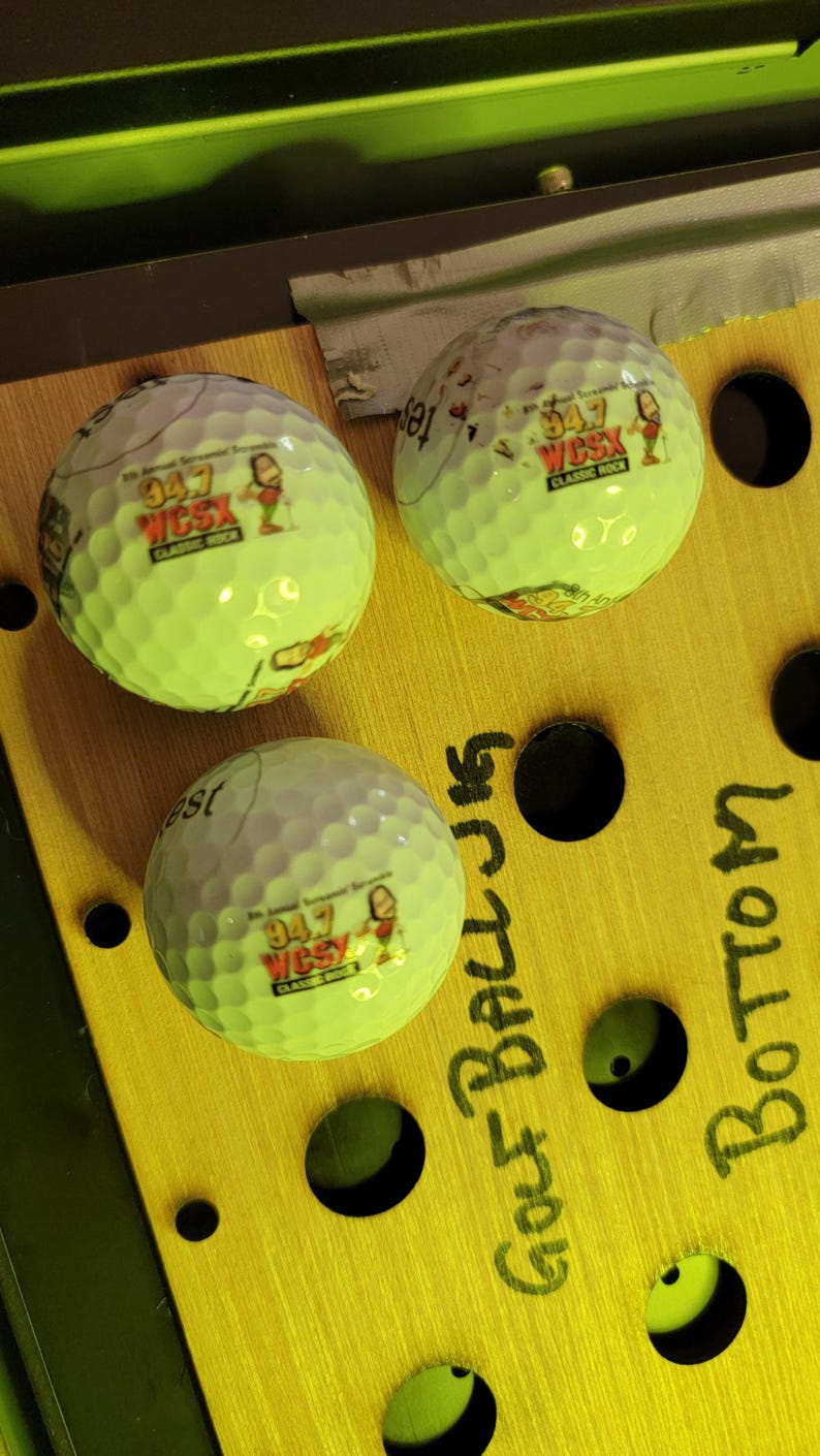 May include: Three white golf balls with dimpled surfaces, each featuring a printed logo that reads 94.7 WCSX Classic Rock with a cartoon character. The balls are positioned on a yellow board with black circular holes and handwritten text.