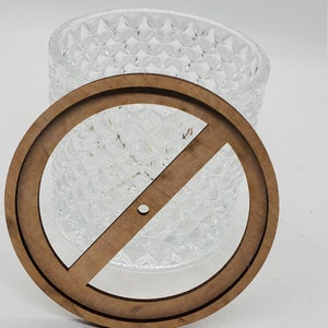 May include: A wooden circle with a diagonal line across it, designed to fit over a glass jar with a textured surface.