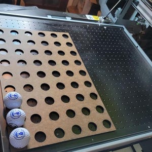 Peut inclure: Gros plan d'une découpeuse laser avec une feuille de bois présentant de multiples découpes circulaires. Trois balles de golf avec un motif bleu et blanc sont placées sur le côté gauche de la feuille.