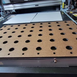 May include: A flatbed industrial printer featuring a brown rectangular vacuum table surface with a grid of circular holes of varying sizes. The machine includes a metallic frame, a print head assembly, and two white rectangular sheets positioned at the top.