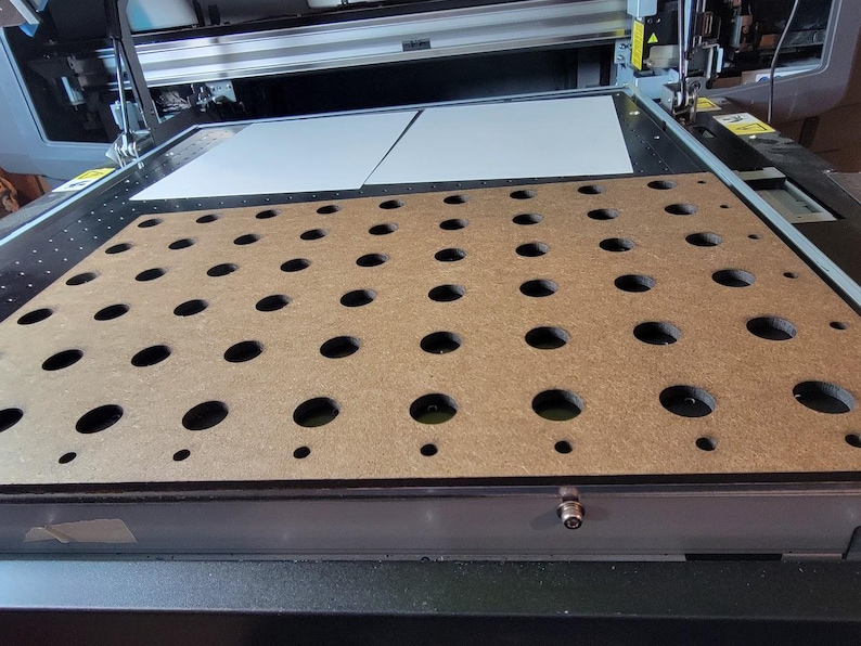 May include: A flatbed industrial printer featuring a brown rectangular vacuum table surface with a grid of circular holes of varying sizes. The machine includes a metallic frame, a print head assembly, and two white rectangular sheets positioned at the top.