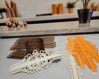 May include: A set of laser cut wooden craft supplies. The set includes two white and two orange decorative pieces with intricate designs, and a brown cardboard base with a grid pattern.