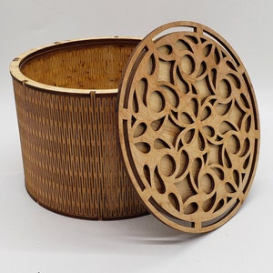 May include: A wooden storage box with a decorative, laser-cut lid. The box is cylindrical with vertical grooves, and the lid features an intricate floral design. The natural wood color gives it a rustic aesthetic.