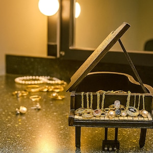 May include: A miniature wooden piano holds an assortment of jewelry, including rings and necklaces. The piano's keys are adorned with various rings, and gold chains hang from the top. Other jewelry pieces, such as a pearl necklace and earrings, are scattered nearby.