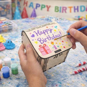 A small wooden craft box is being painted with bright birthday designs, showing how easy and fun these boxes are for kids to decorate. The smooth wood surface and simple shape make them perfect for creative projects, party activities, and personalized gifts that feel handmade and special.