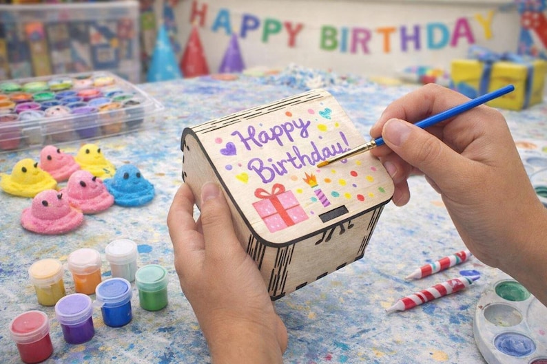 A small wooden craft box is being painted with bright birthday designs, showing how easy and fun these boxes are for kids to decorate. The smooth wood surface and simple shape make them perfect for creative projects, party activities, and personalized gifts that feel handmade and special.