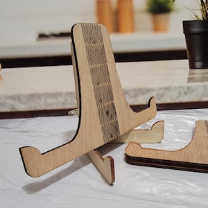 Puede incluir: Un soporte de teléfono de madera con un diseño decorativo cortado con láser. El soporte está hecho de madera marrón claro y tiene un diseño simple y moderno.