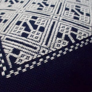 Könnte beinhalten: Ein dunkel marineblaues Stoffquadrat mit komplexer weißer geometrischer Stickerei. Das Design besteht aus sich wiederholenden Rauten- und Kreuzmustern, die in einem dichten, linearen Stil gestickt sind und einen scharfen Kontrast zum dunklen Hintergrund bilden.