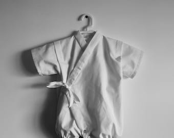 May include: A white baby kimono with a tie closure and button closures at the bottom. The kimono is hanging on a white hanger against a white wall.