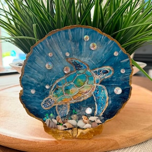 Op de afbeelding: Een decoratieve schelp beschilderd met een levendige blauwe zeeschildpad die in diepblauw water zwemt. De schelp is voorzien van iriserende ronde accenten, goudkleurige randen en een verzameling kleine, natuurlijke schelpjes aan de onderkant.