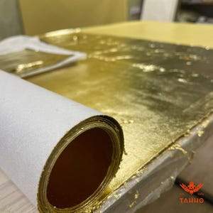 May include: A roll of gold leaf paper with a white backing is partially unrolled, revealing sheets of gold leaf. The gold leaf covers a surface, with a brush and more gold leaf visible in the background. The image is well-lit, highlighting the metallic sheen.