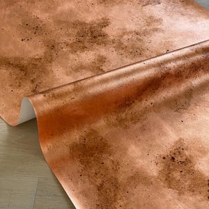 May include: A roll of copper-colored wallpaper with a metallic sheen. The paper has a mottled pattern with darker brown spots and streaks, creating a textured effect. The roll is partially unrolled, revealing the white backing.