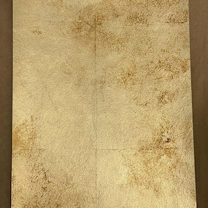 May include: A rectangular sheet featuring a metallic gold finish with a distressed, textured surface. The material displays variations in tone, ranging from light champagne gold to deeper, burnished bronze patches across the entire flat surface.