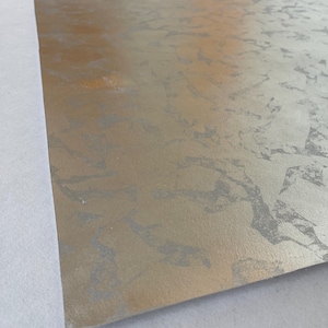 May include: A roll of metallic wallpaper with a textured, mottled silver and gold finish. The paper has a subtle sheen and a pattern that resembles aged metal or stone. The roll is partially unrolled, revealing the paper's surface.