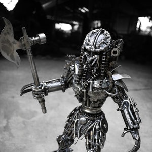 May include: A metal sculpture of a Predator character from the movie franchise. The sculpture is made of various metal parts, including nuts, bolts, and gears. The Predator is holding a metal axe in its right hand. The sculpture is standing on a metal base.
