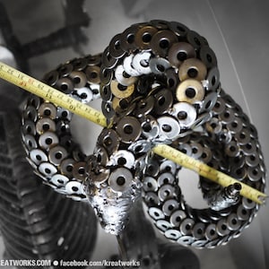 May include: A metal sculpture of a snake made from hundreds of silver and gold washers. The snake is coiled and measures approximately 91 cm in length.