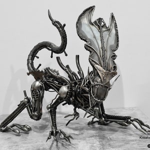 May include: A detailed, silver-colored metal sculpture of an alien creature with a long tail and a large head. The sculpture is made of recycled metal parts and is a unique piece of art.